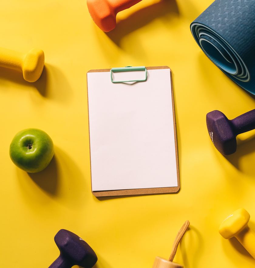 Dumbbells, a yoga mat, a green apple and a water bottle around a clipboard showing a sample daily plan